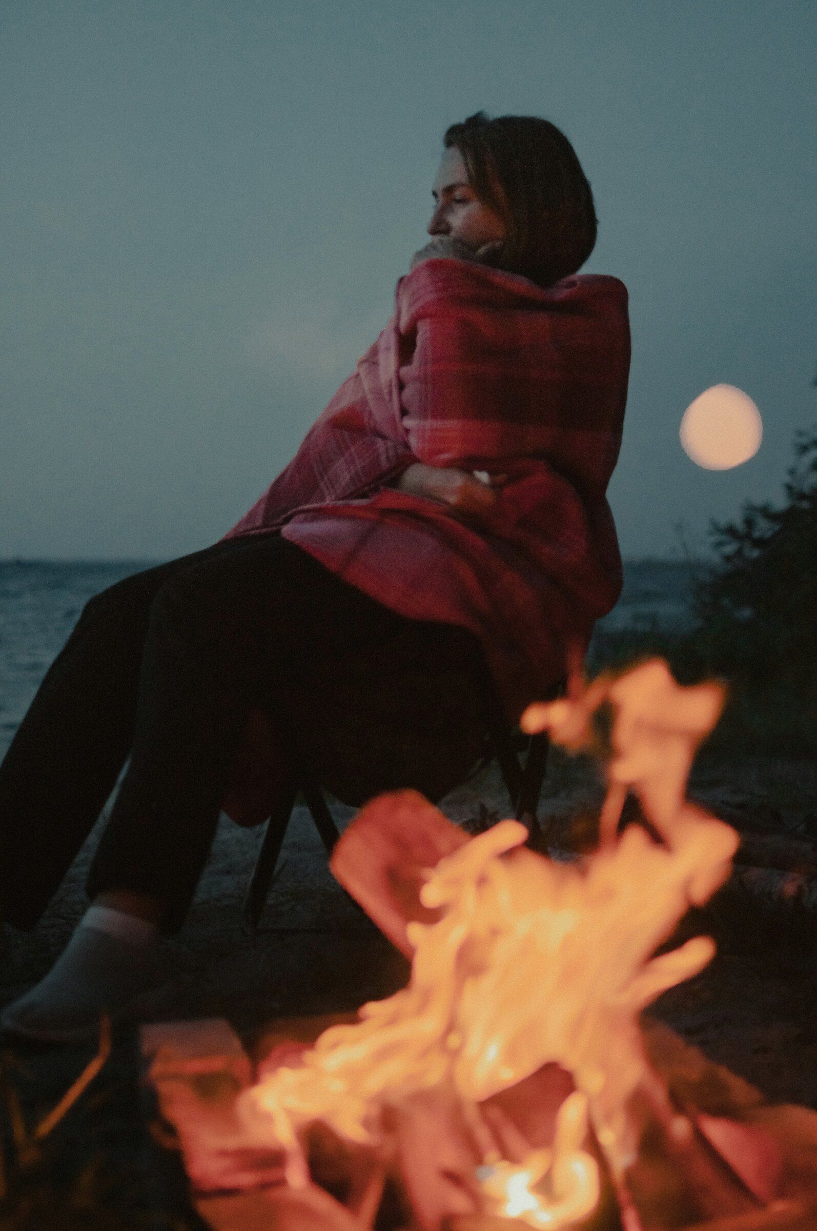 A woman wrapped in a plaid blanket beside a fire under a rising moon