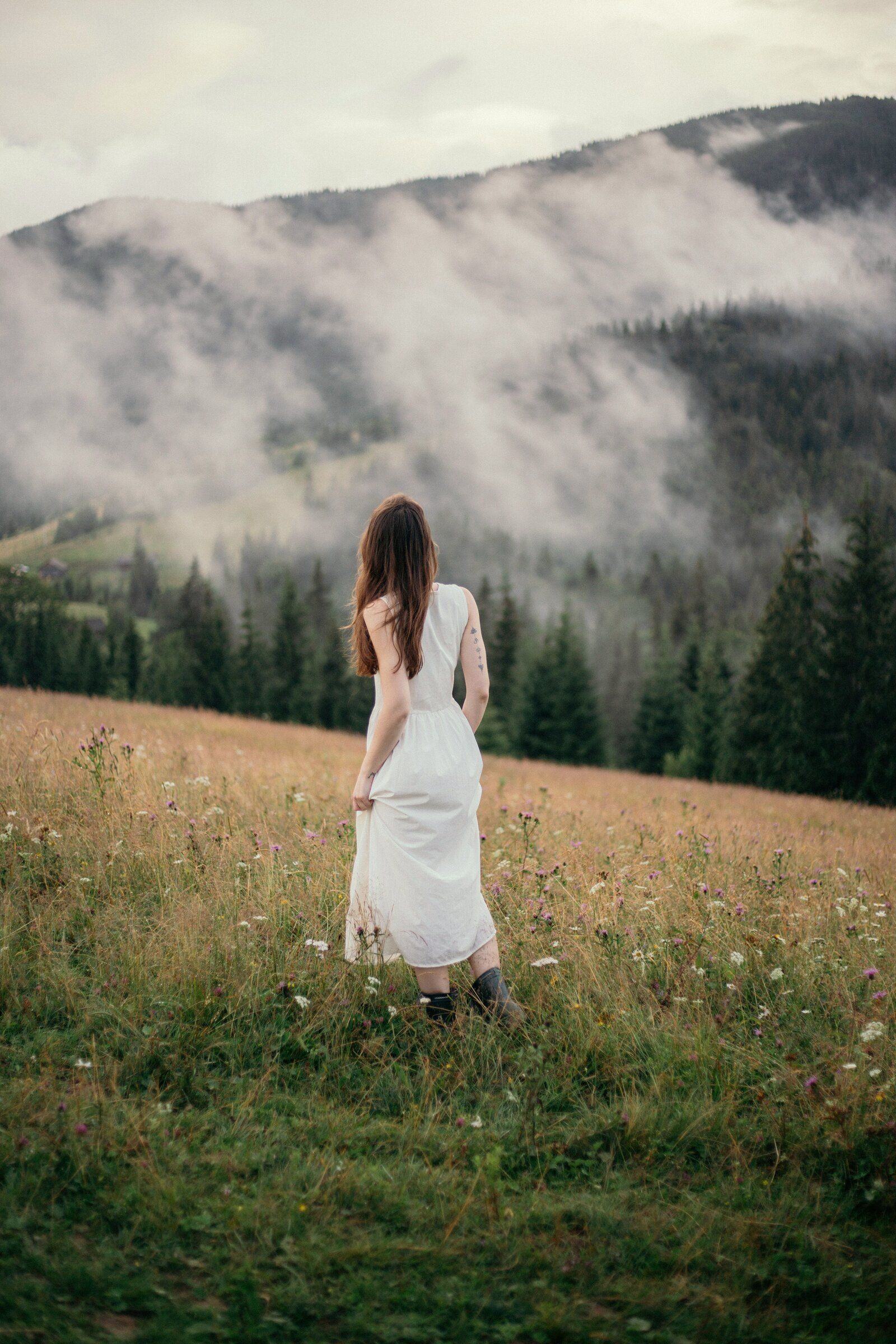 A woman in a white dress walking into a wildflower meadow below misty mountains