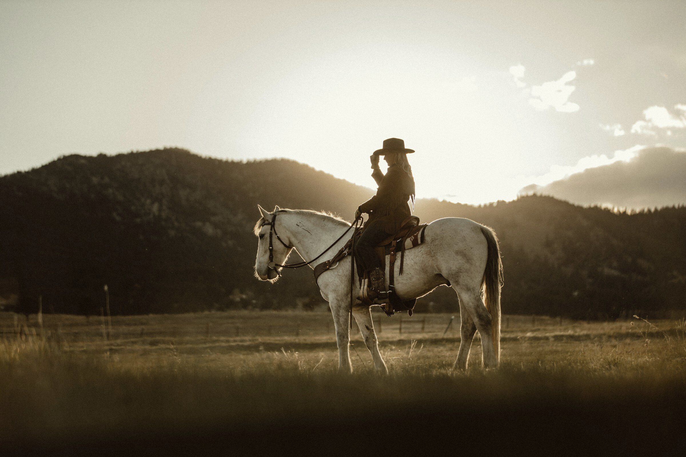 A woman in a cowboy hat on a white horse against the mountains at sunset